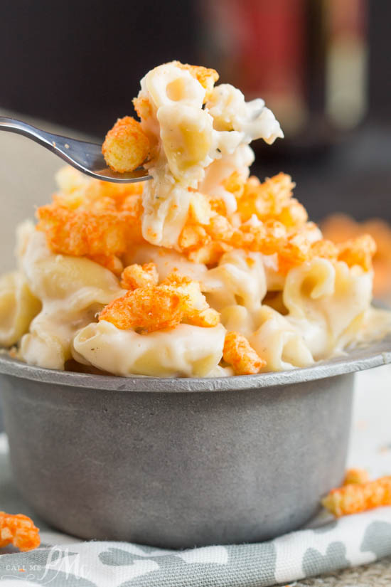 A fork holds a bite of cheese pasta, topped with crushed Cheetos, above a metal cup.
