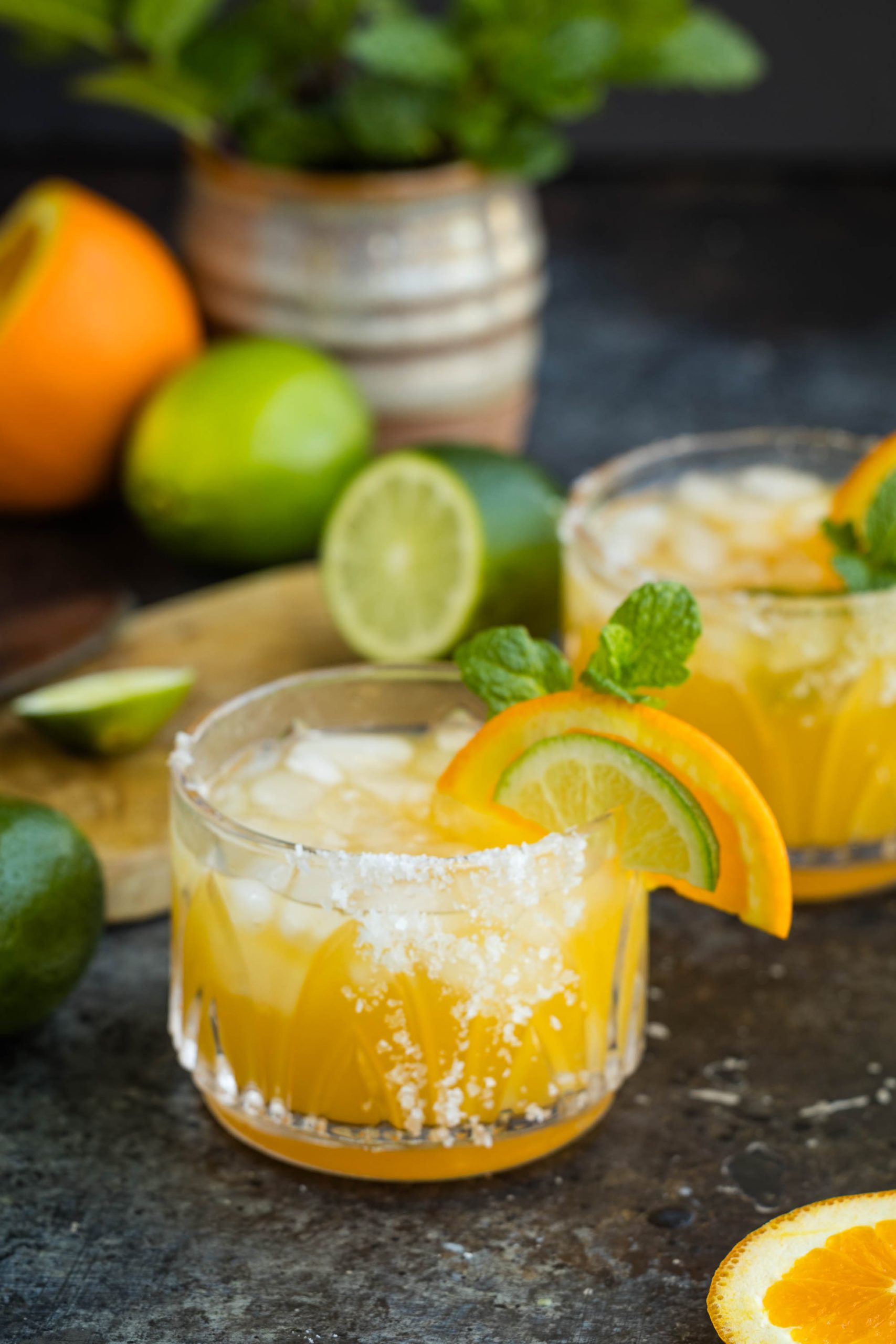 Two glasses of Italian Amaretto Margarita garnished with lime and orange slices on a dark table, with fresh fruits and mint in the background.