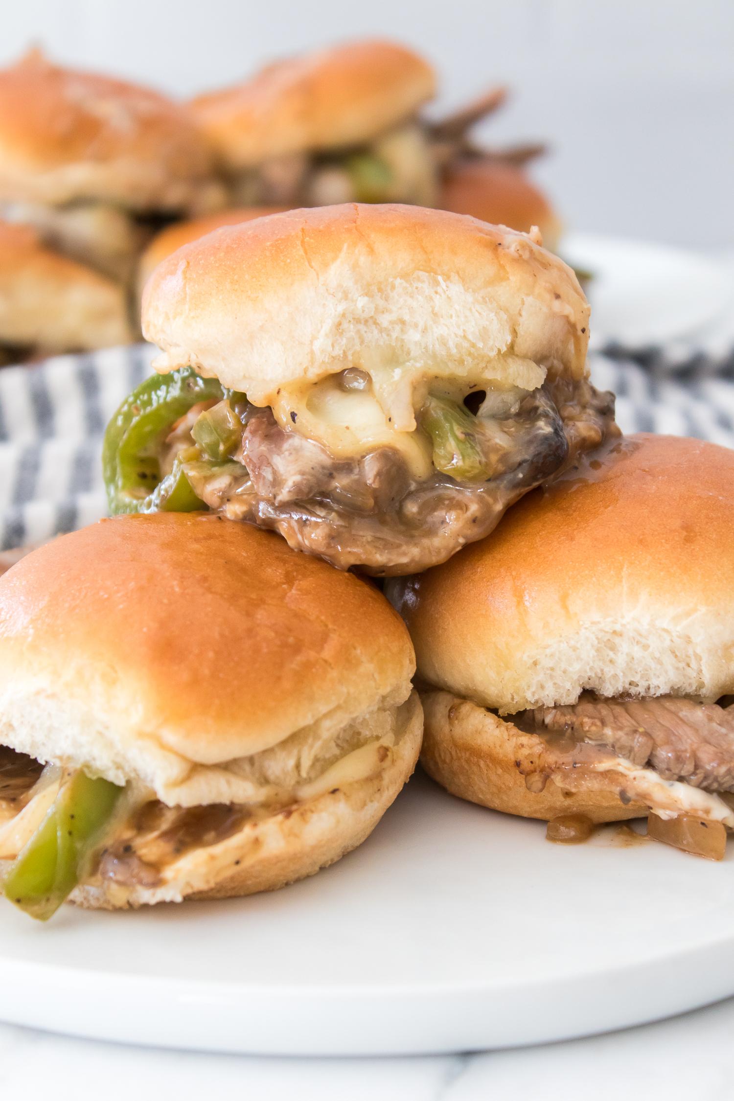 Three Philly Cheesesteak Sliders with beef, onions, peppers, and cheese on buns are stacked on a white plate. A striped cloth is in the background.