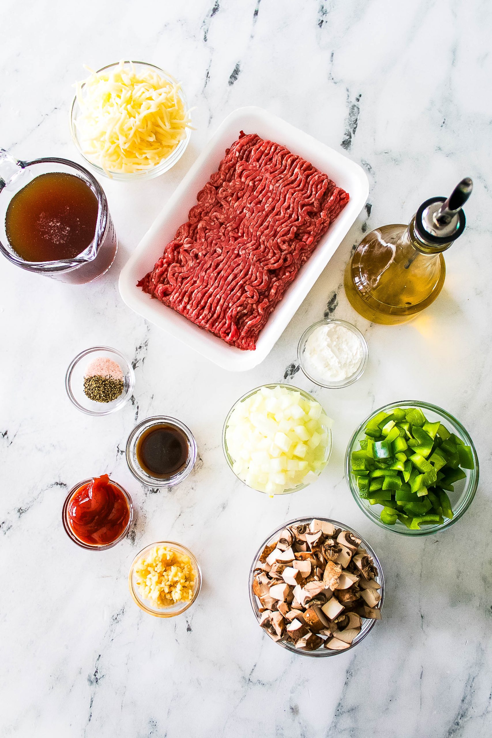 Ingredients on a marble surface: ground beef, shredded cheese, olive oil, beef broth, diced onions, green peppers, chopped mushrooms, minced garlic, ketchup, seasoning, and Worcestershire sauce.